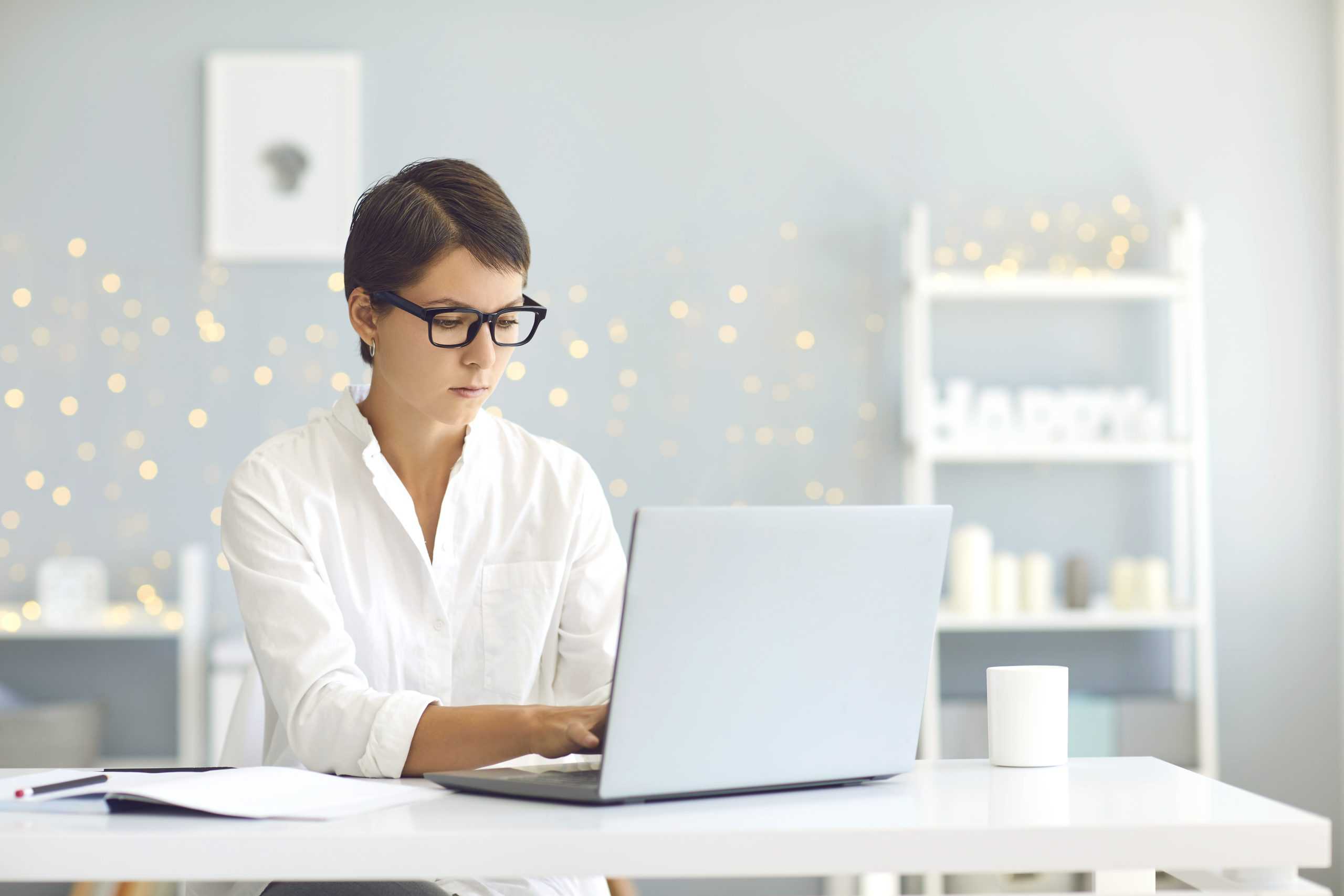 Freelance, distant working, online communication, business concept. Young business lady in glasses stting and working on laptop online from home with cosy room interior at background